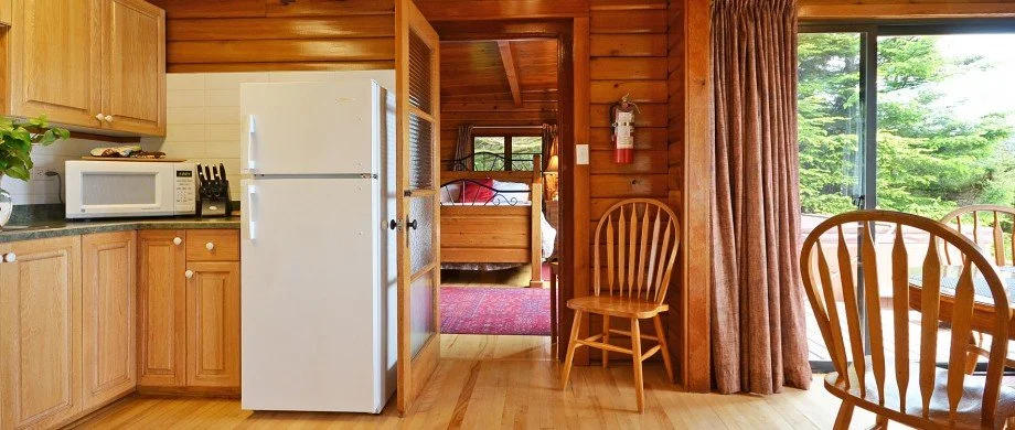 Interior view of a cozy wooden cabin with a kitchen and dining area, showing a white refrigerator, microwave, wooden chairs, a glass sliding door with curtains, and a glimpse of a bedroom in the background.