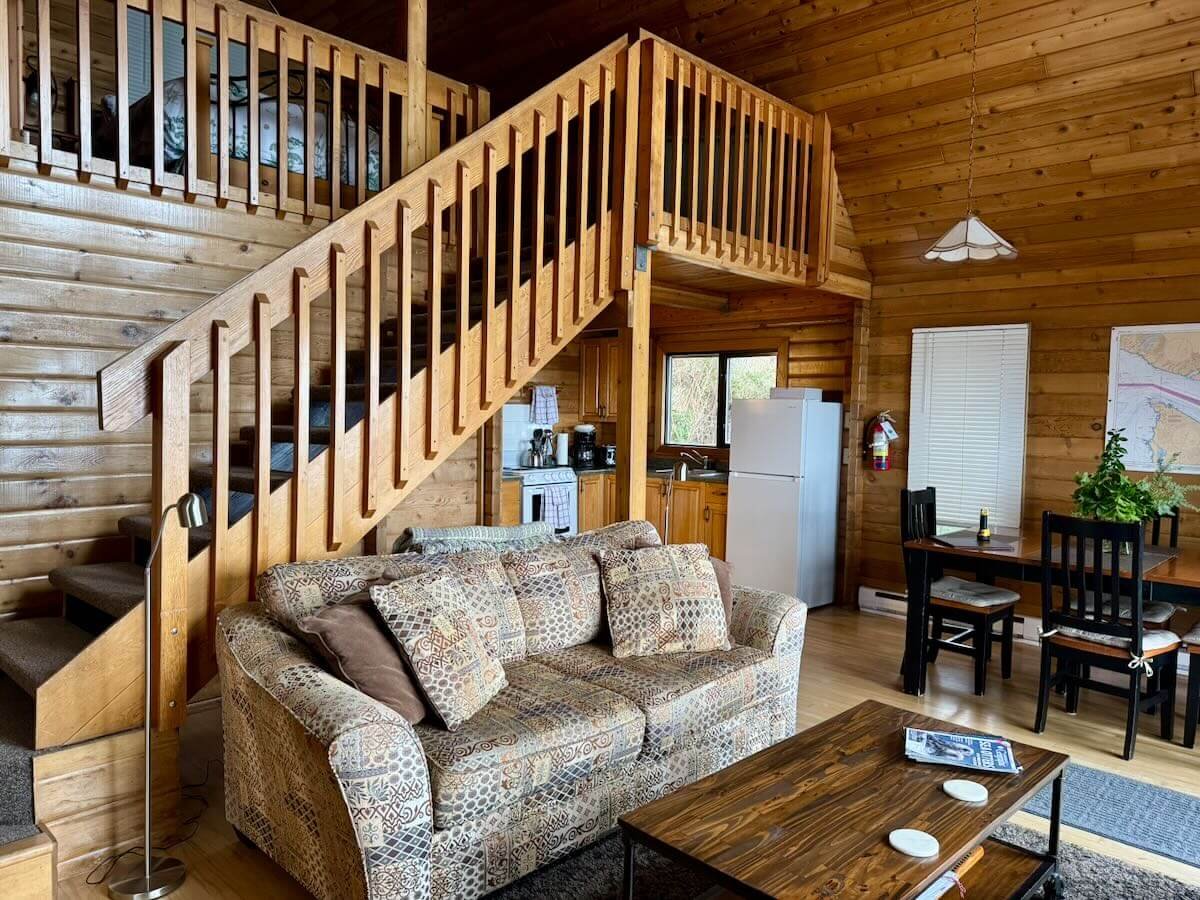 Cabin A living room with a sofa, coffee table, and a staircase to bedroom loft. A dining table with chairs, and kitchen are in the background.