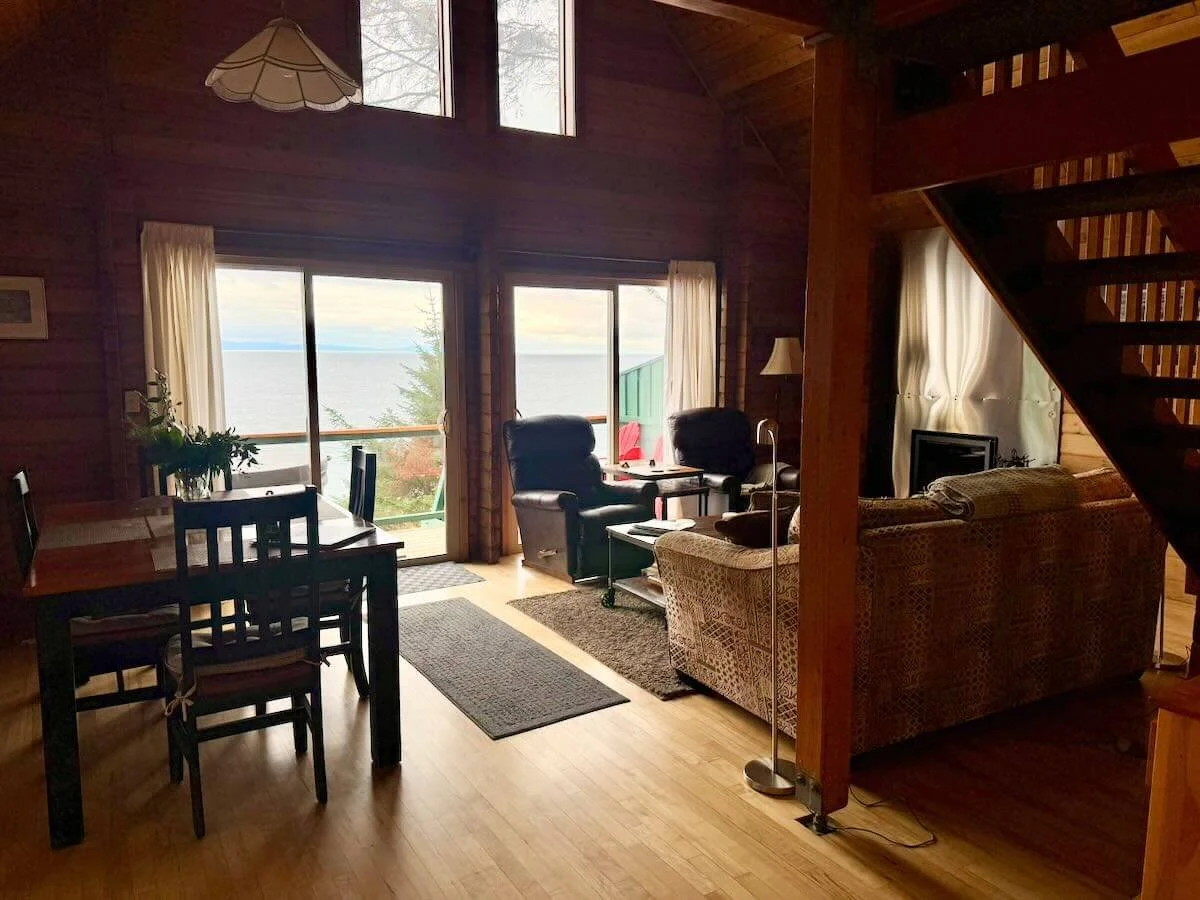 Cabin A interior with wooden walls, a dining table, and a living area with two armchairs and a sofa, with large windows and glass doors overlooking the ocean.