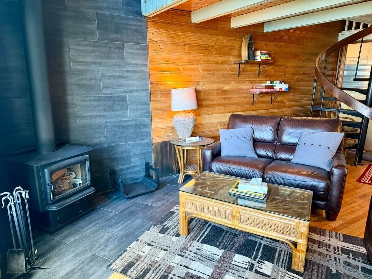 Cabin 12 living area with a wood and gray tiled wall, a dark brown leather sofa, a rattan coffee table, a floor lamp next to a rattan side table, and wooden shelves on the wall. There is a spiral staircase on the right and a wood stove on the left.
