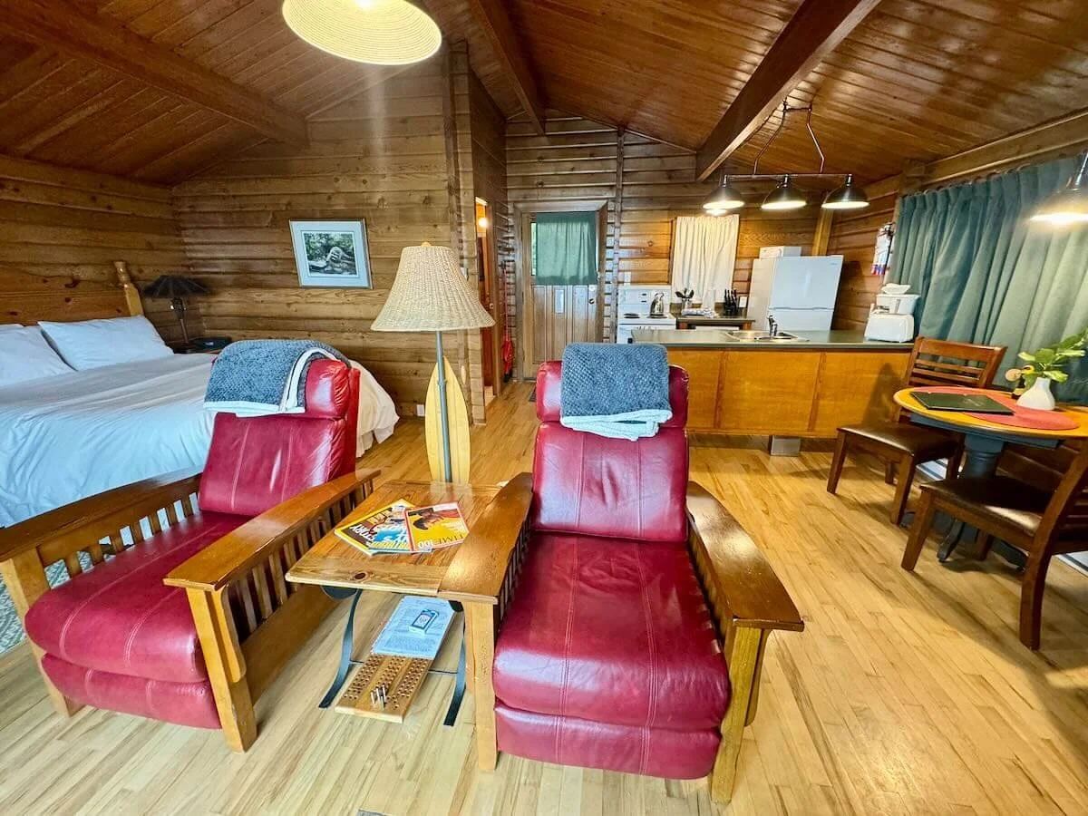 Interior of Cabin B with a kitchenette, a bed with white linens, a small table with magazines, red leather chairs with blue towels, a floor lamp, and a dining table with chairs. The kitchen area has white appliances and pendant lighting.