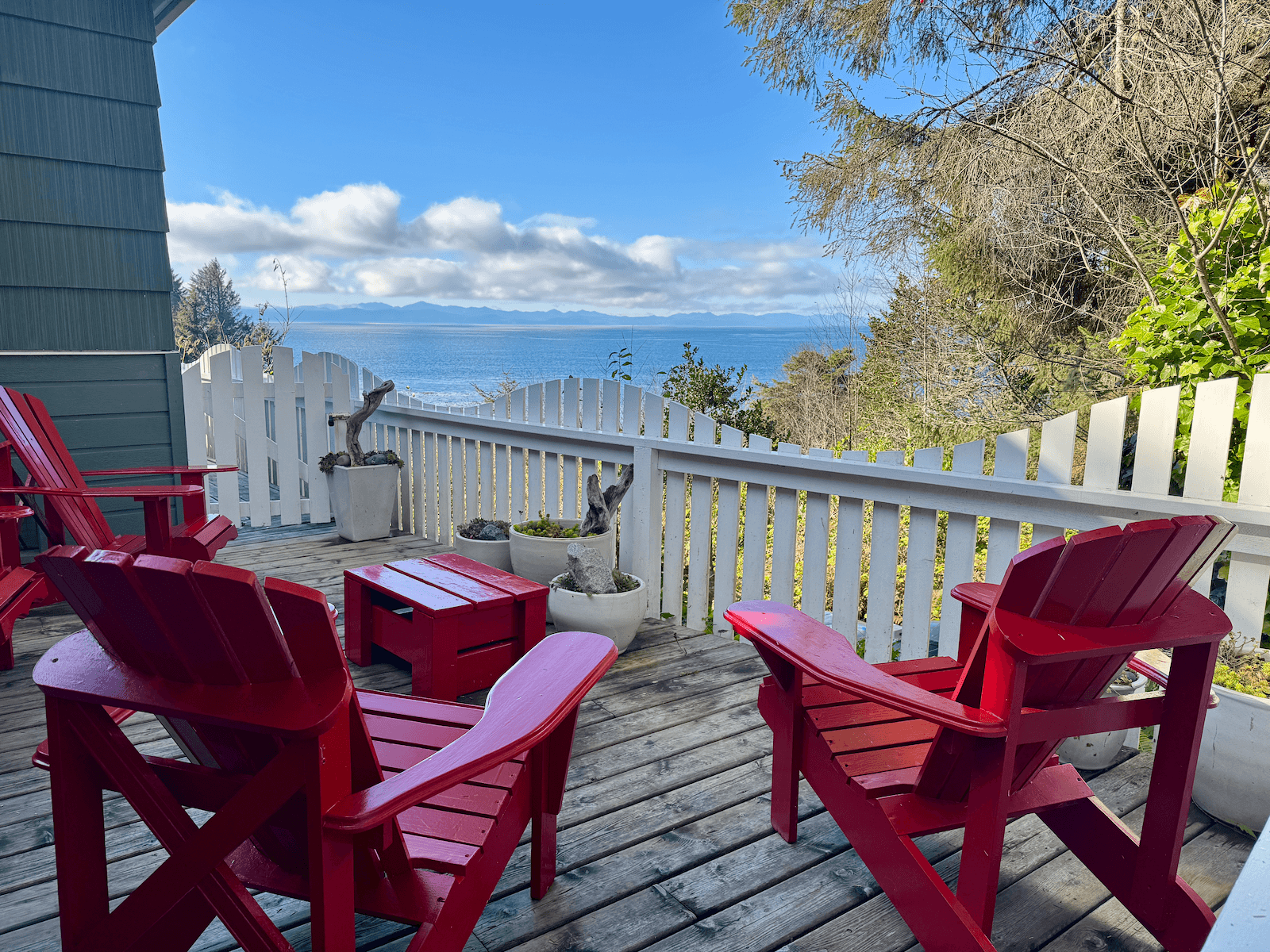 A balcony with red Adirondack chairs, potted plants, and a view of the ocean with mountains in the distance under a partly cloudy sky.