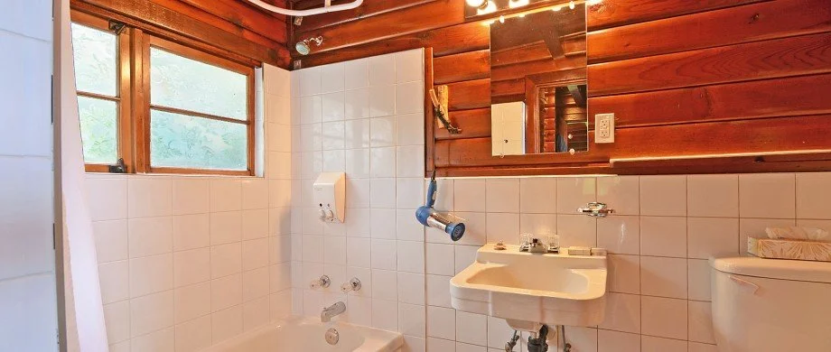 Bathroom with wooden walls, a window, a white bathtub, a small sink, a mirror, and a toilet.