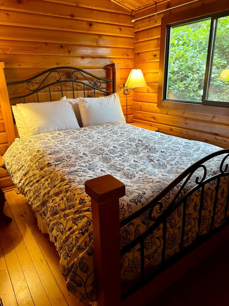 A cozy bedroom with wooden walls, a metal bed frame with white pillows, a floral bedspread, a wooden bedside table, and a window showing greenery outside, with a yellow wall-mounted lamp beside the bed.