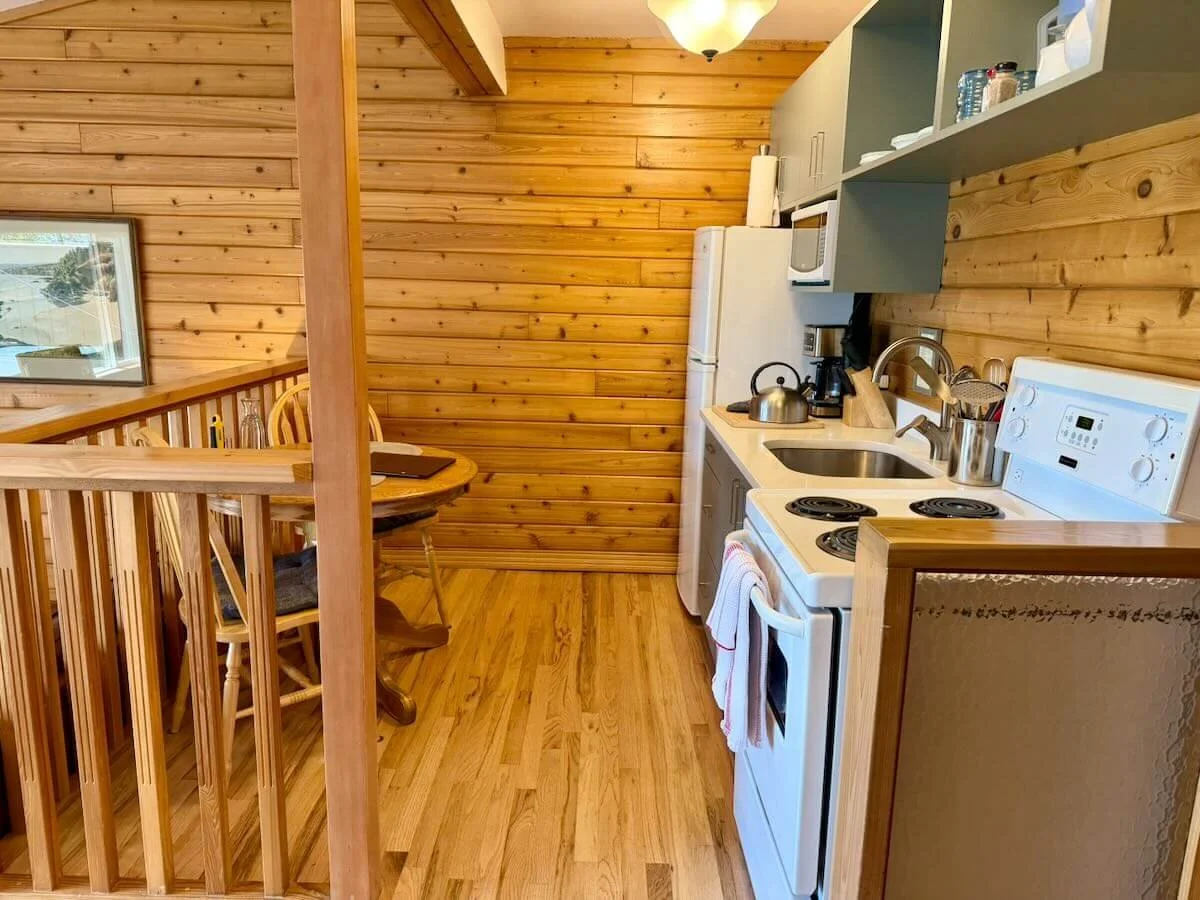 A small kitchen with wooden walls and flooring, featuring white appliances including a refrigerator, stove, and microwave. There is a sink, kettle, coffee maker, and some utensils on the counter. A dining area with a wooden table and chairs is visible to the left.