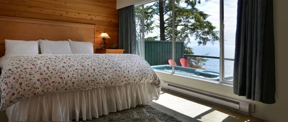 Bedroom with a large bed, floral bedding, wooden wall, bedside lamp, and a view of a balcony with trees and water outside the window