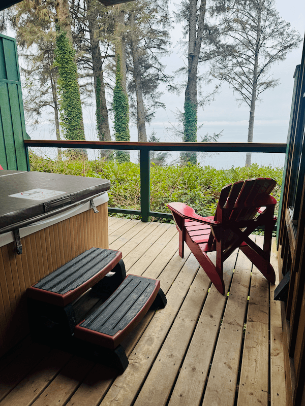 A wooden deck with a hot tub, a red Adirondack chair, and a black step stool in front of a green railing. The scene overlooks a forest with tall trees and a body of water in the distance.
