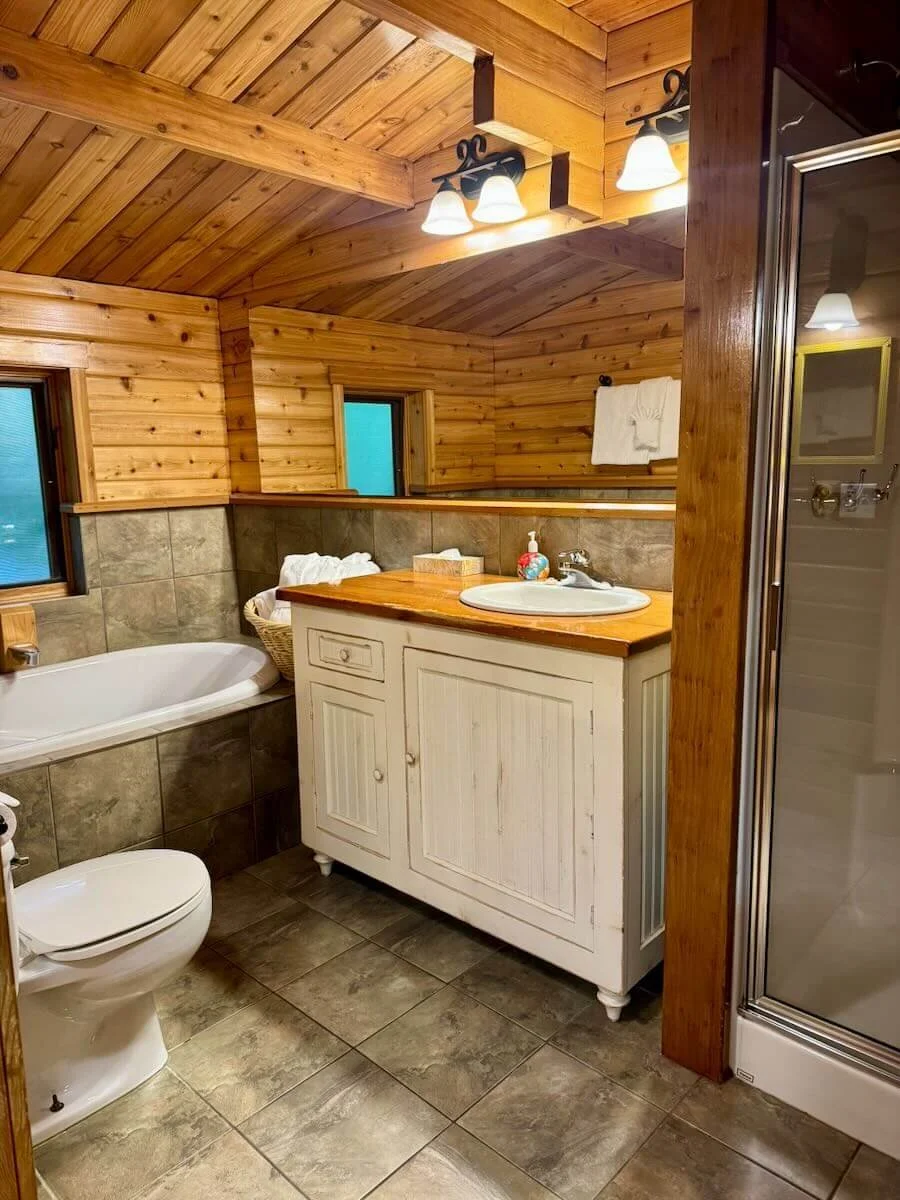 Bathroom with wood-paneled walls and ceiling, containing a corner bathtub, a white vanity with a wooden countertop, a sink, and a shower stall. There are small windows with frosted glass, and warm lighting fixtures above the mirror.