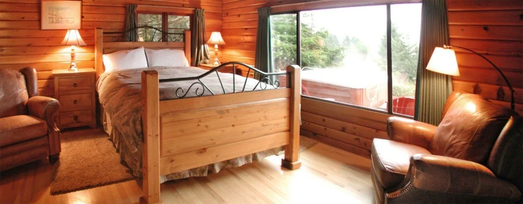 Cozy cabin bedroom with wooden walls, a large bed with white linens, two nightstands with lamps, a leather armchair, a sofa, and large windows showing greenery outside.