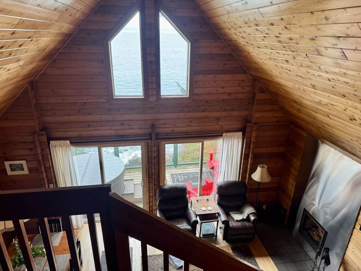 Cabin A living room with large windows, a stone fireplace, leather armchairs, and a balcony with red chairs overlooking the ocean.