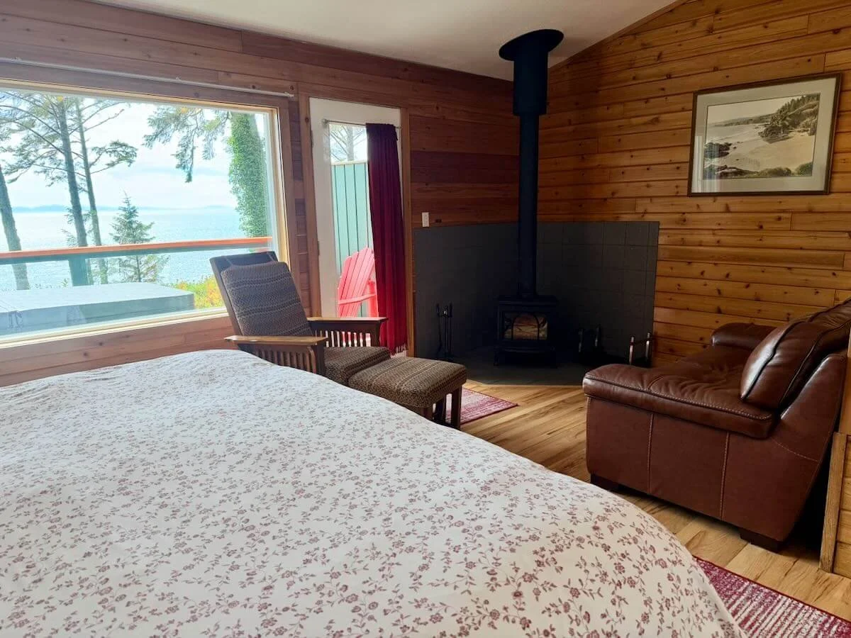 Interior of a cozy bedroom with wood-paneled walls, large window showing trees and water, a bed with floral bedding, a rocking chair with ottoman, leather armchair, and a fireplace in the corner.