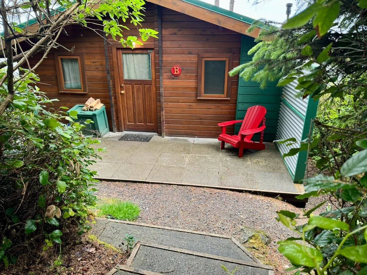 Exterior of Cabin B - Raccoon with a green and brown exterior, a wooden door, two windows, and a red Adirondack chair on a concrete porch, surrounded by greenery.