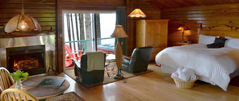 Cozy cabin bedroom with a fireplace, leather chairs, a glass door leading to a balcony with outdoor seating, and a neatly made bed with a basket of towels.