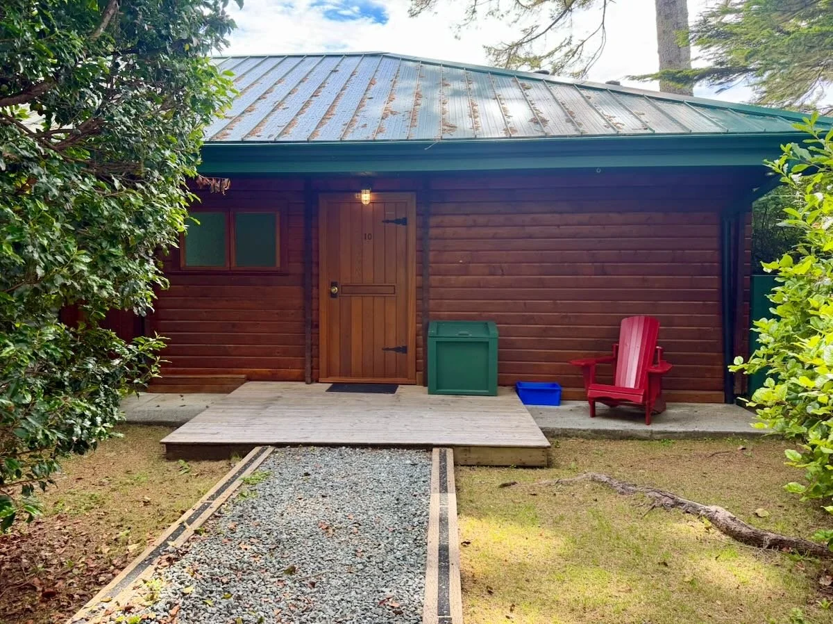 Cabin 10 exterior with a sloped metal roof, a wooden door, a window, a red Adirondack chair, and a gravel walkway with one step leading to the entrance.