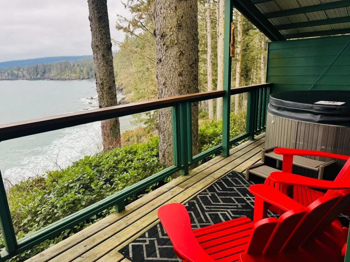 A balcony with red Adirondack chairs, a small table, a hot tub, and a view of trees and water.