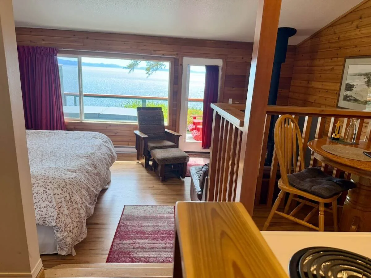 Cozy hotel room with large window showcasing a view of a lake, bedroom with bed and patterned covers, small sitting area with chairs, and a wooden dining table with chairs.