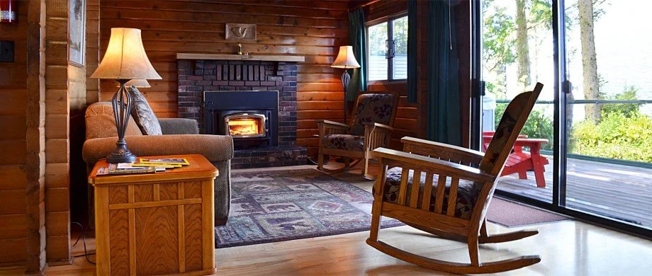 Cozy living room with a wood-burning fireplace, wooden walls, a gray sofa with pillows, a wooden side table with magazines, two lamps, an armchair, a rocking chair, and sliding glass doors leading to an outdoor deck with a red bench and greenery outside.
