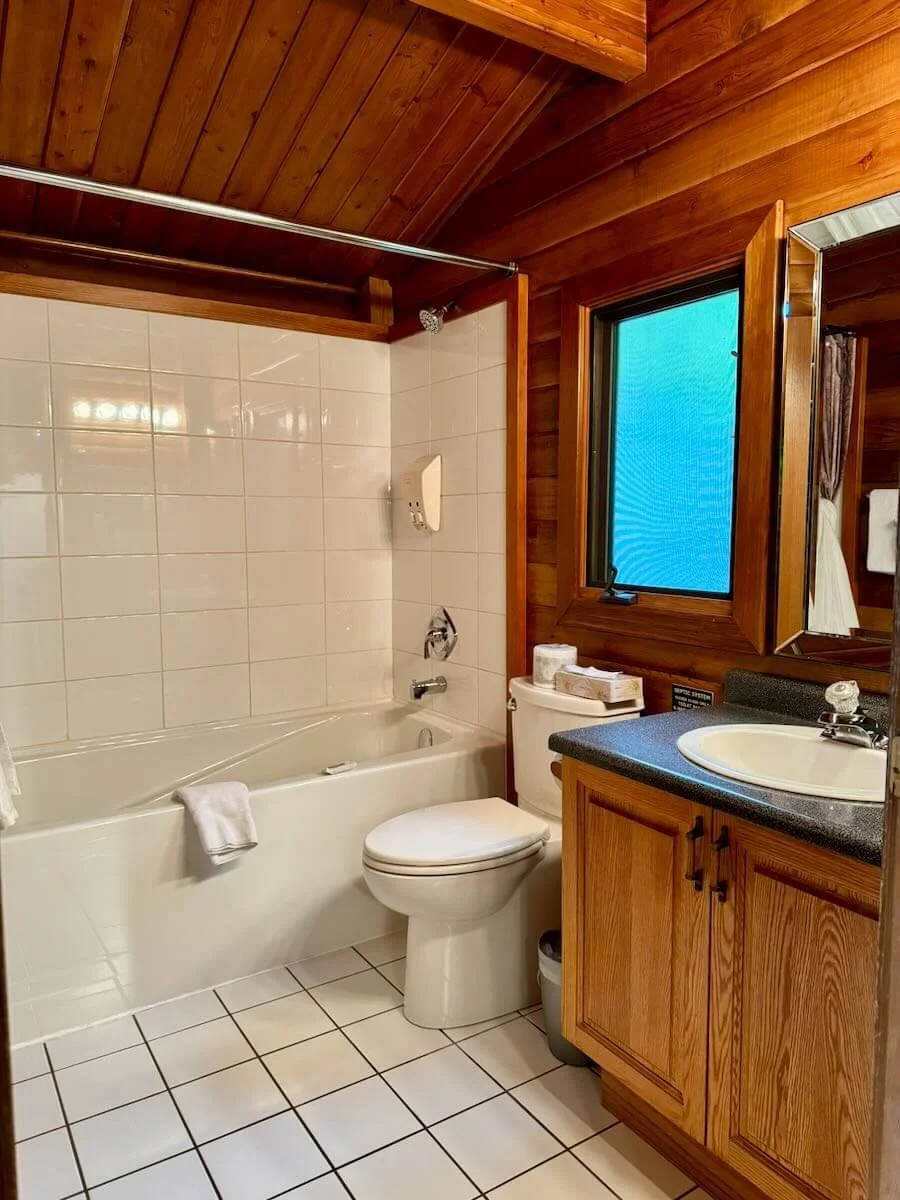 Cabin B bathroom with wooden-paneled ceiling and walls, white tile floor, white bathtub with a towel, toilet, and a wooden vanity with a sink, mirror, and window.