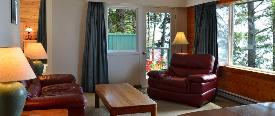 Living room with two red leather sofas, a wooden coffee table, two table lamps, large windows showing a forest view, and a door leading outside.