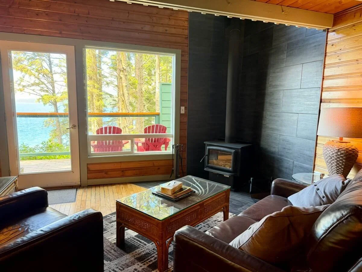 Cozy living room with wooden walls, a fireplace with a fire, two leather armchairs, a glass coffee table, and a window with a view of trees outside.