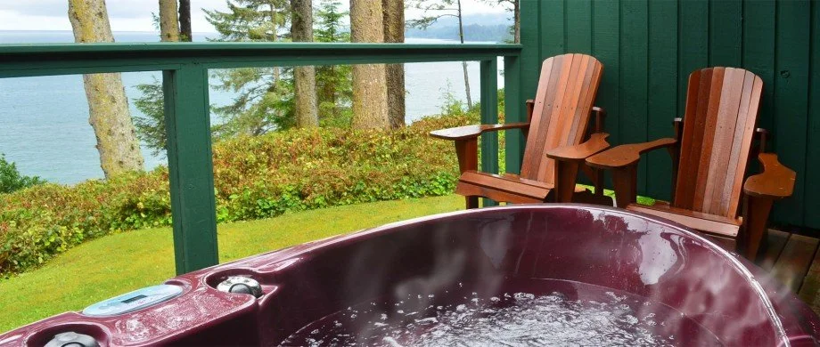 A deck with two wooden chairs overlooking a view of trees and water, with a hot tub in the foreground.