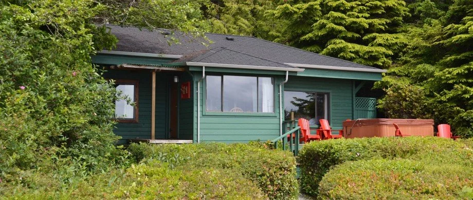 A green house with a dark roof surrounded by lush greenery, red chairs, and a hot tub on the porch.