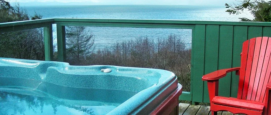 Hot tub on a wooden deck overlooking a body of water with a green railing and a red Adirondack chair.