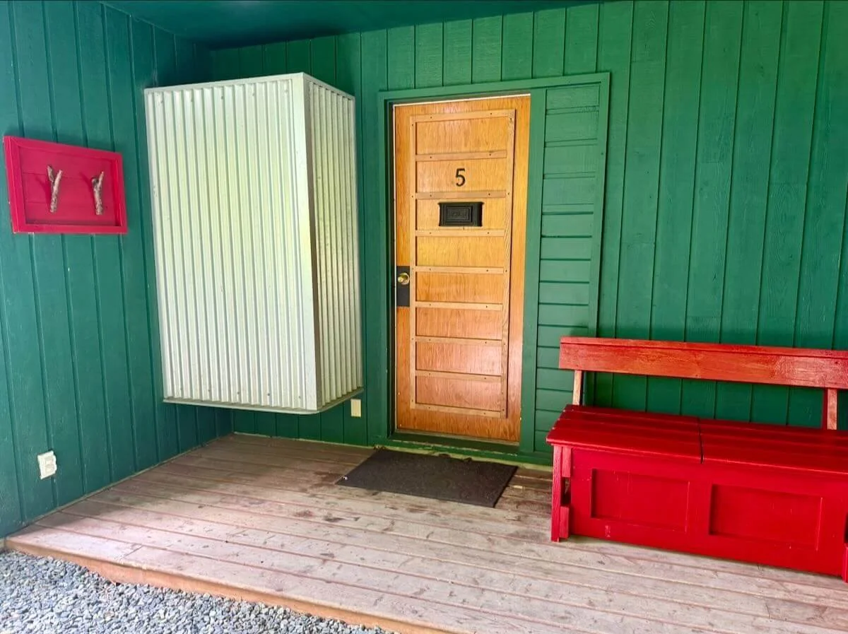 Green porch with wooden door marked '5', red wooden bench, small red wall feature with eagle hooks, white metal utility box, black doormat, gravel ground.