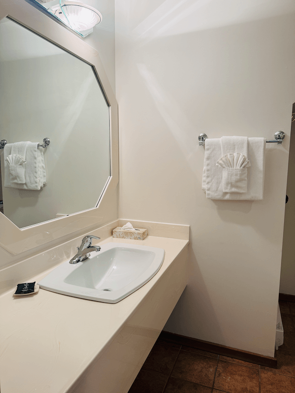 Bathroom sink with a large mirror above, a soap dish, and a towel rack with folded towels. Well-lit with light-colored walls and tiled floor.