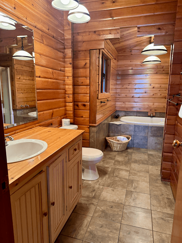 A rustic bathroom with wooden paneled walls and ceiling, a mirror over a wooden vanity with a white sink, a toilet, and a bathtub. There is a basket and towels near the bathtub, with tile flooring and a small window on the wall.
