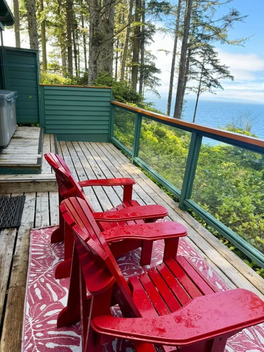 Cabin 12 hot tub and two red Adirondack chairs on a wooden deck, overlooking trees and a body of water in the distance.