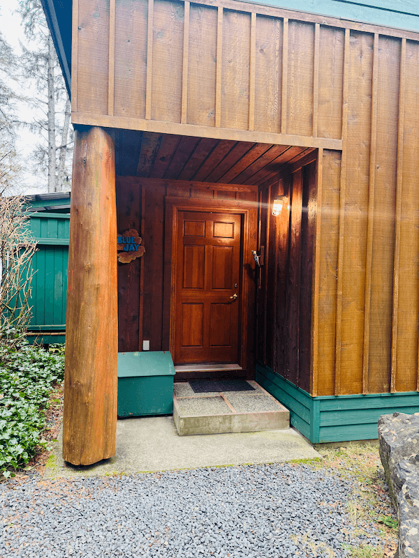 Cabin Blue Jay entrance with a dark wood door, a porch light, and a small set of concrete stairs. A green mailbox and decorative sign are nearby, with greenery and trees in the background.