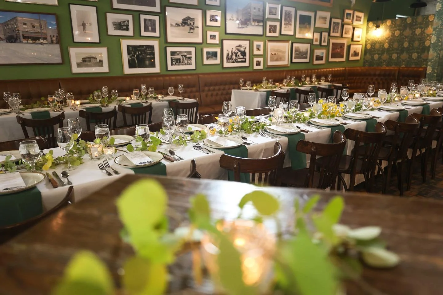 A restaurant dining room set up for a large event, with long tables decorated with green garlands and white tablecloths, arranged with plates, silverware, wine glasses, and candles, against a wall decorated with framed black-and-white photographs.