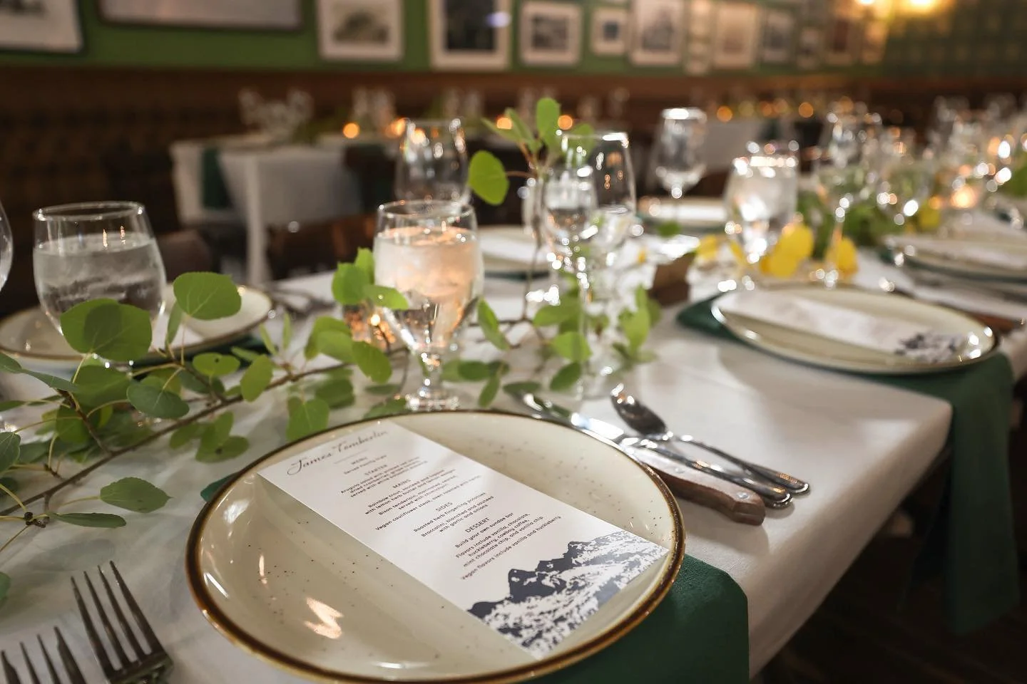 A decorated table set for a formal event with white tablecloth, green napkins, plates with dinnerware, and glasses of water and champagne, adorned with green foliage and small yellow flowers.