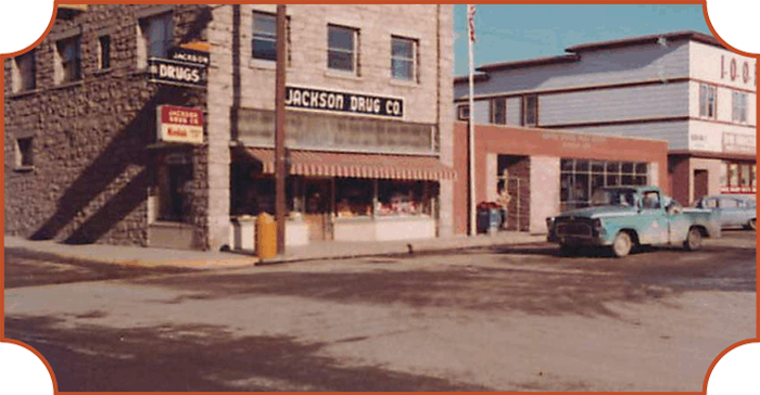 Street view of Jackson Drug Co, a historic storefront with a brick facade and awning, parked cars nearby, and an adjacent building with a fire escape.