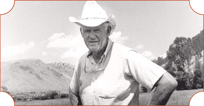 A black and white photo of an older man wearing a cowboy hat, glasses hanging from his shirt, and a light-colored shirt. He is outdoors with hills, trees, and a partly cloudy sky in the background.
