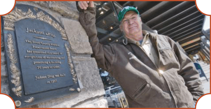 A man in a green cap and brown jacket standing next to a historical marker on Jackson Drive, smiling and leaning against a stone wall.