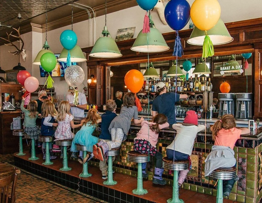 Children sitting on bar stools at a bar counter with balloons hanging from the ceiling, with bartenders preparing drinks in the background.