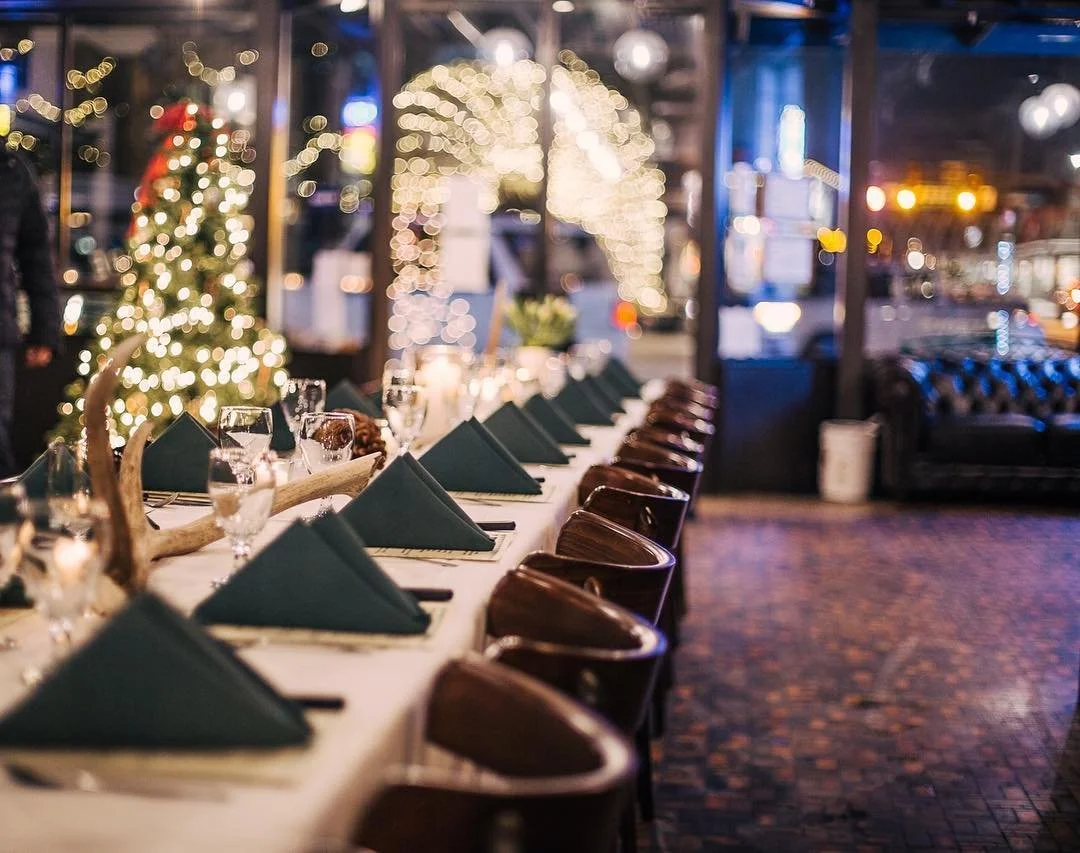A long dining table set for a holiday celebration with green napkins, wine glasses, and a centerpiece featuring antlers. In the background, blurred Christmas trees with lights and festive decorations are visible, creating a cozy holiday atmosphere.