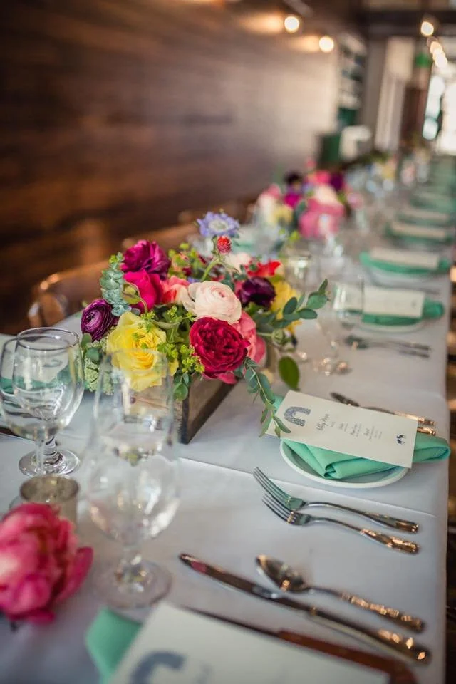 A long dining table decorated with a flower centerpiece of pink, purple, yellow, and white flowers with green leaves. Place settings include green napkins, wine glasses, water glasses, plates, and silverware, in a warmly lit room with wooden walls.
