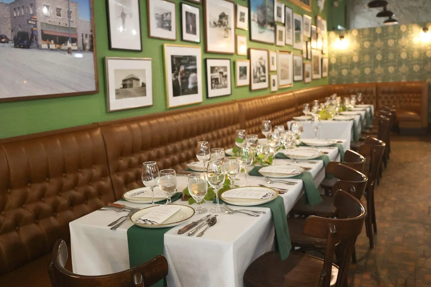 A neatly set dining table in a restaurant with white tablecloths, green napkins, and glassware. The restaurant's wall is decorated with numerous framed black-and-white and color photographs.