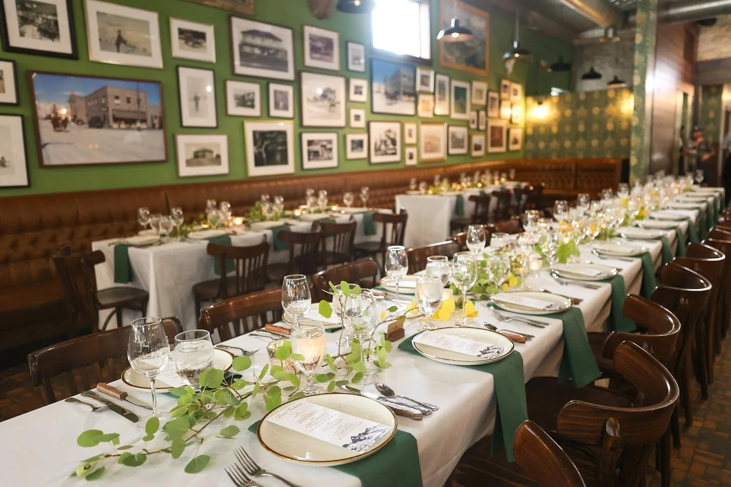 Elegant dining room with long tables set for a special event, decorated with white tablecloths, green napkins, glassware, plates, and leafy centerpieces, with a green wall adorned with black-and-white photographs.