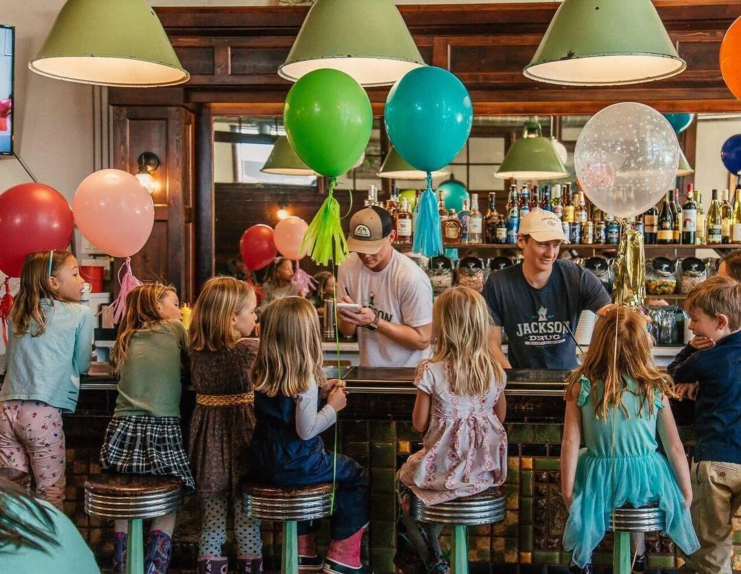 Children celebrating birthday party at a bar or restaurant with balloons, sitting on bar stools, with two young men behind the bar.