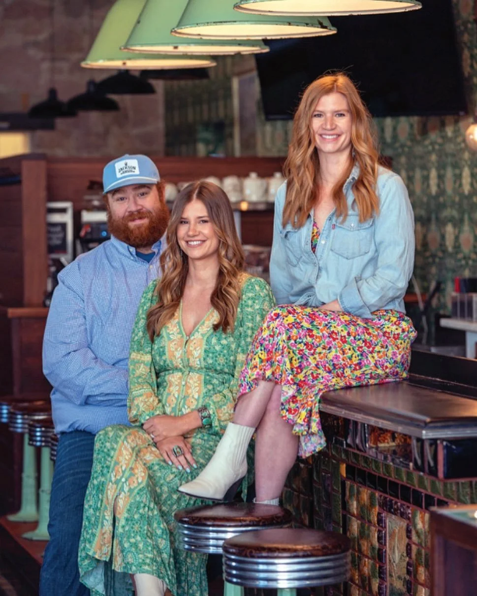 Three people, two women and one man, smiling in a cozy restaurant or bar. One woman with red hair sitting on a barstool, wearing a colorful floral skirt and a light denim shirt. The other woman with brown hair in a green dress, sitting on a stool. The man with a beard and mustache, wearing a blue shirt and a cap, standing behind them.