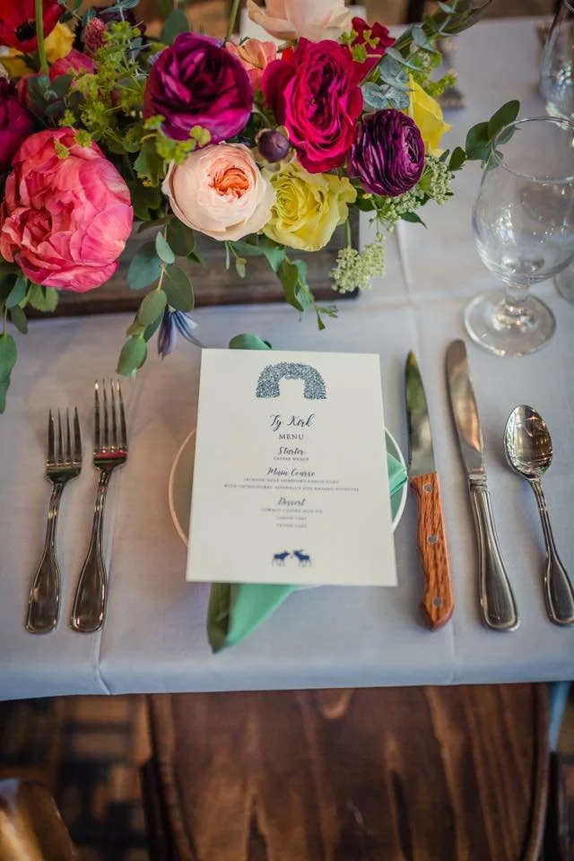 A dining table setting with a large bouquet of pink, purple, yellow, and peach flowers, a menu card, silverware, a water glass, and a green napkin, with wooden flooring visible.
