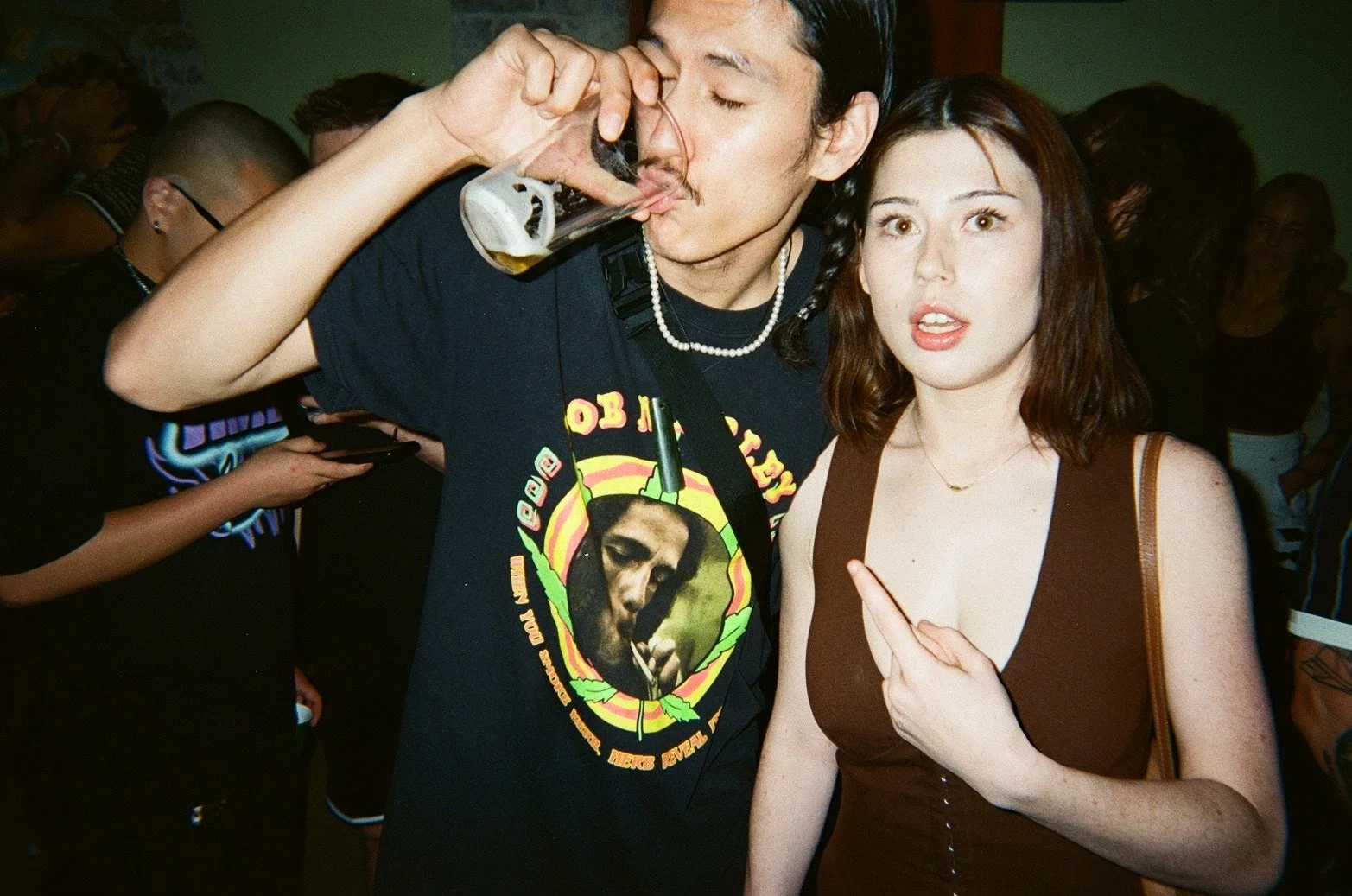 A man with long dark hair wearing a black t-shirt with a Bob Marley graphic and a pearl necklace is drinking from a glass of beer. Next to him is a woman with shoulder-length red hair wearing a brown top, pointing at herself, with a crowd of people i