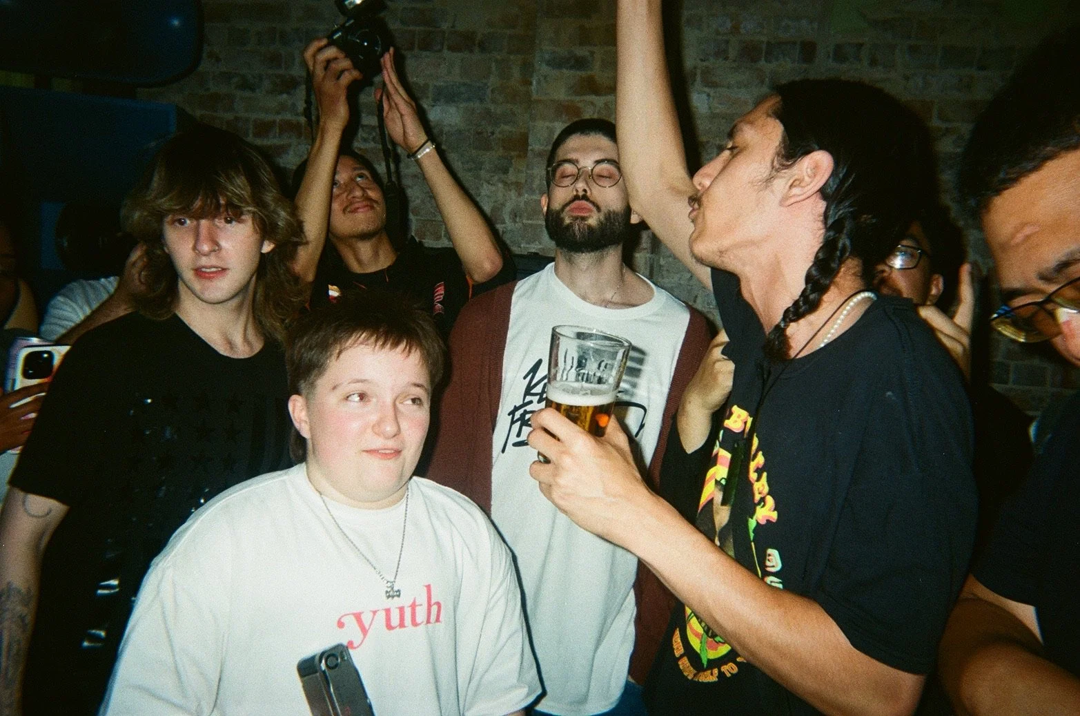 Group of young adults at a party, some holding drinks, enjoying socializing against a brick wall background, with some taking photos and engaging with each other.
