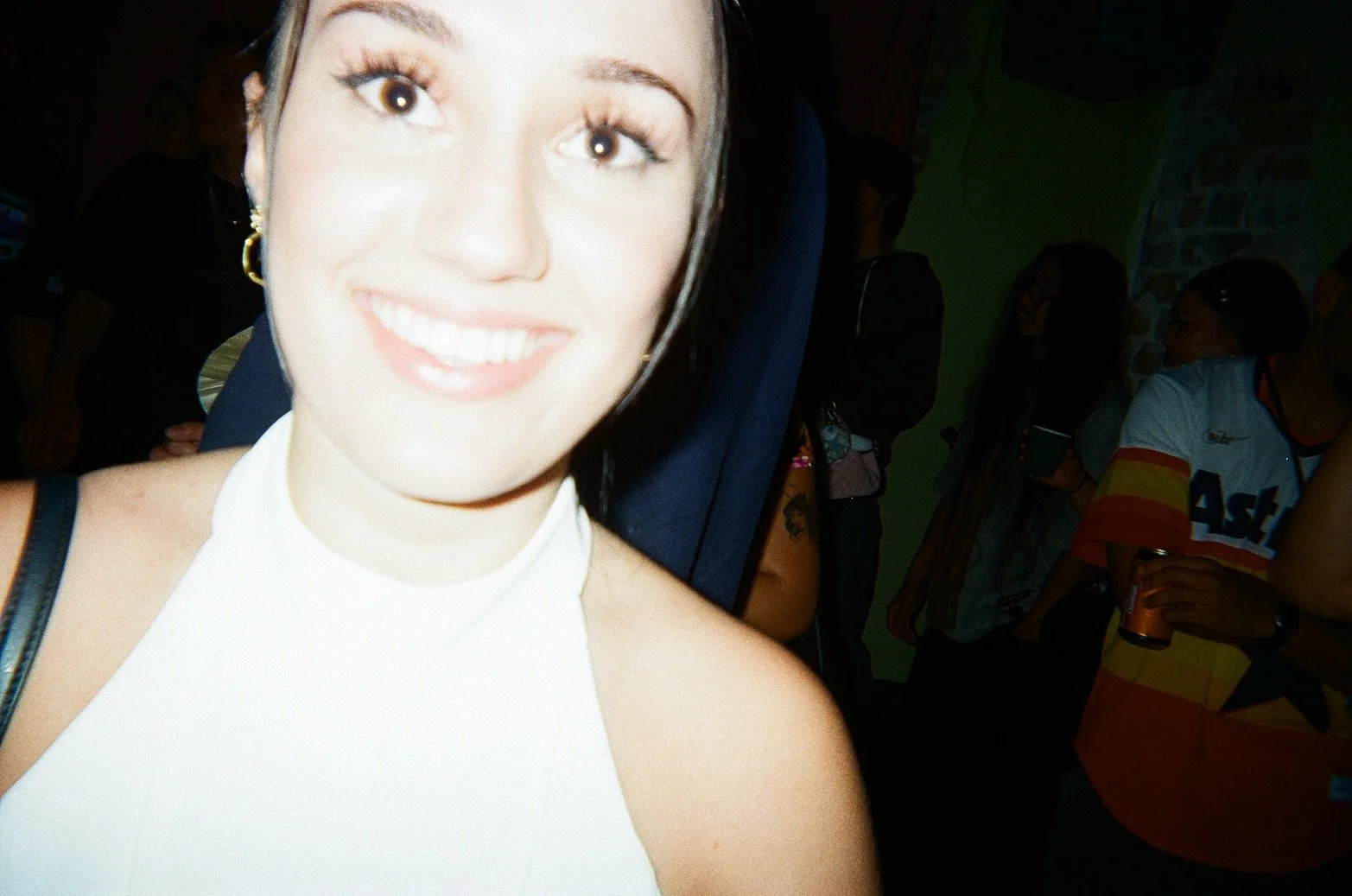 Close-up of a woman smiling with a bright white top, gold earrings, and dark hair, at a social gathering with other people in the background.