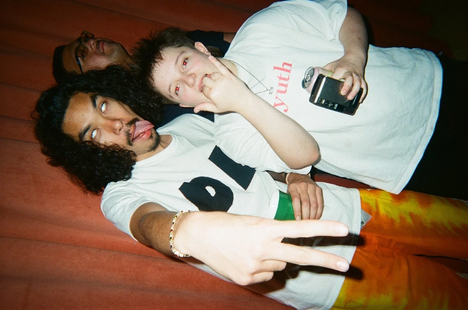 Three young men lying on a wooden floor, posing playfully for the camera. One in the foreground is making a peace sign with his fingers, sticking out his tongue, and has curly hair. The second, in the middle, has short dark hair, and is holding a can