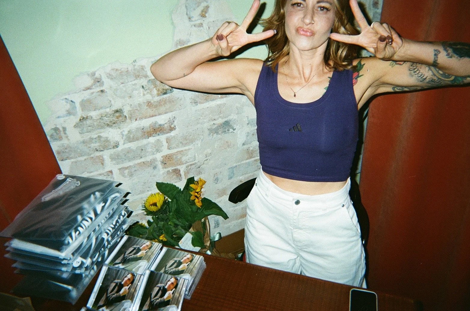 A woman making a peace sign with both hands, wearing a blue crop top and white pants, standing behind a desk with brochures and a small vase of sunflowers, against a backdrop of a brick wall with a green-painted area.